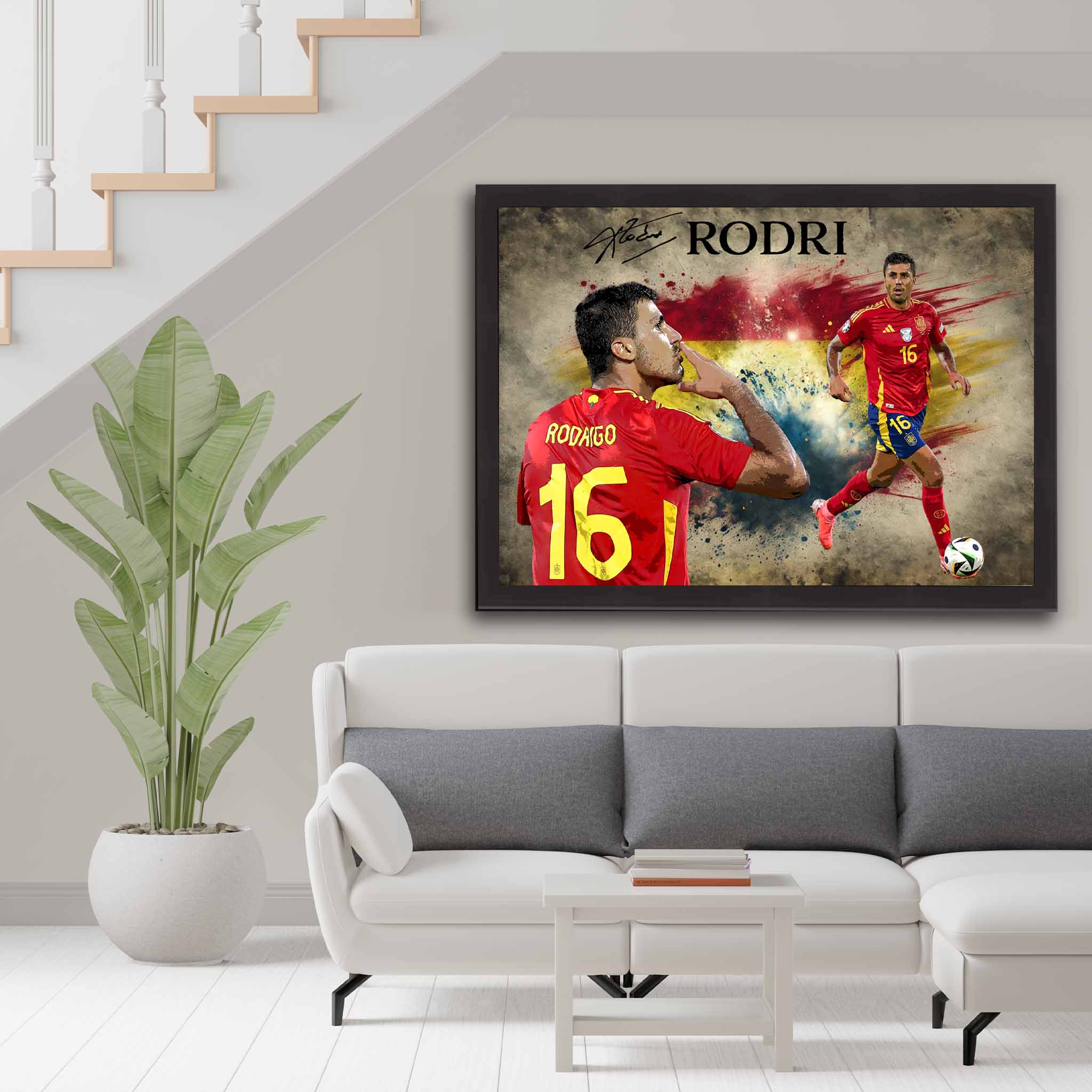 Framed Rodri Spain Soccer Art poster hanging on a modern living room wall above a white sofa with grey cushions, next to a green plant. The poster features Rodri in his red Spain jersey, celebrating a goal and in action on the field. The background has the Spanish flag with grunge textures and vibrant colors.